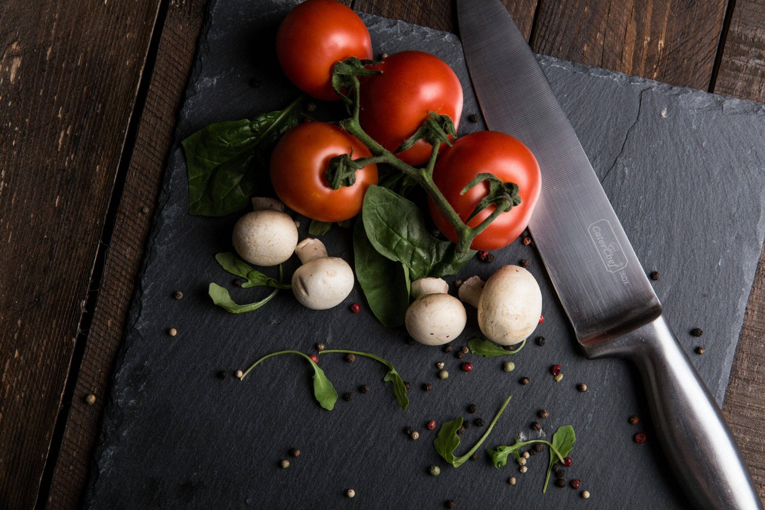 Fresh tomatoes on the vine, white mushrooms, spinach leaves, and scattered peppercorns on a dark slate cutting board with a chef’s knife.

